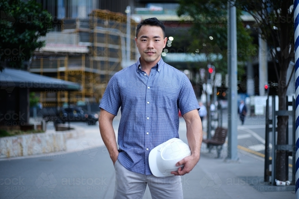 Asian man holding hard hat with city background - Australian Stock Image