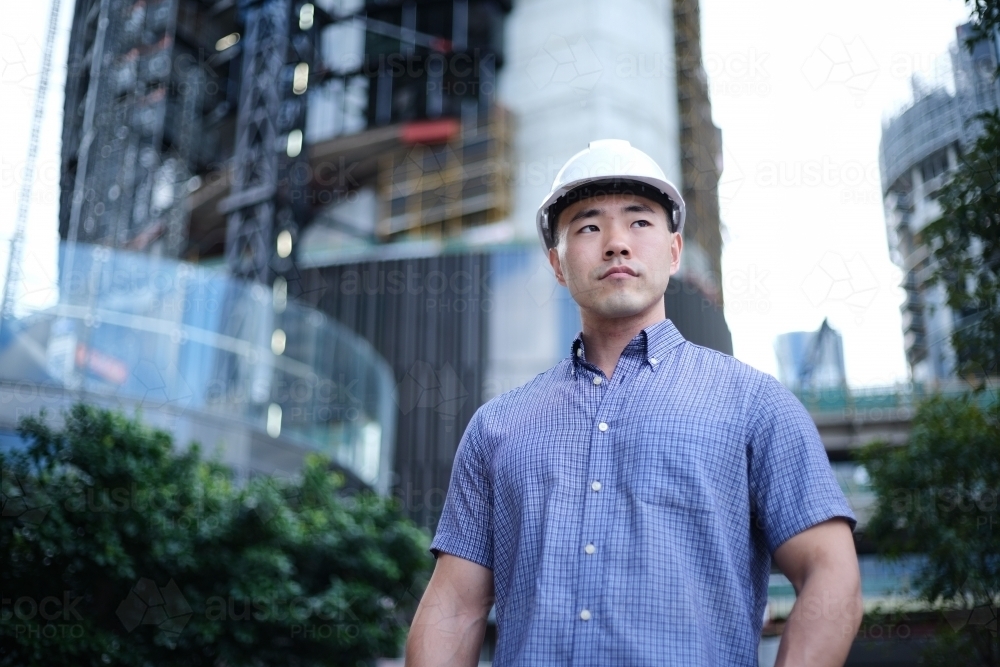 Asian man wearing hard hat with city background - Australian Stock Image