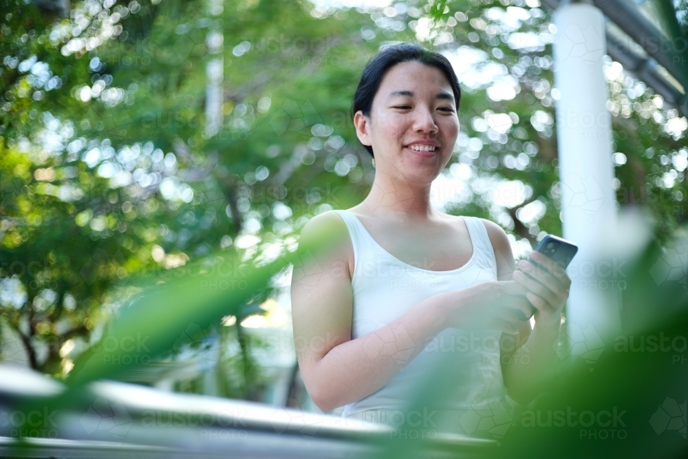 Asian woman on her phone can be seen through the green foliage - Australian Stock Image