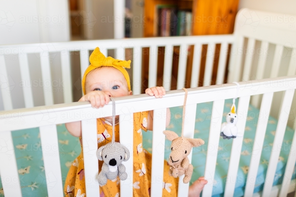 Aussie baby girl in cot pulling up on edge of bed with hanging toys - Australian Stock Image