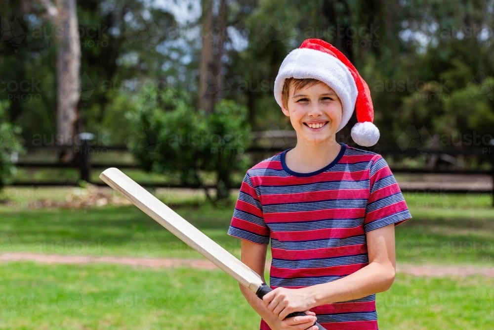 Aussie boy ready to play backyard cricket in front yard at Christmastime wearing santa hat - Australian Stock Image