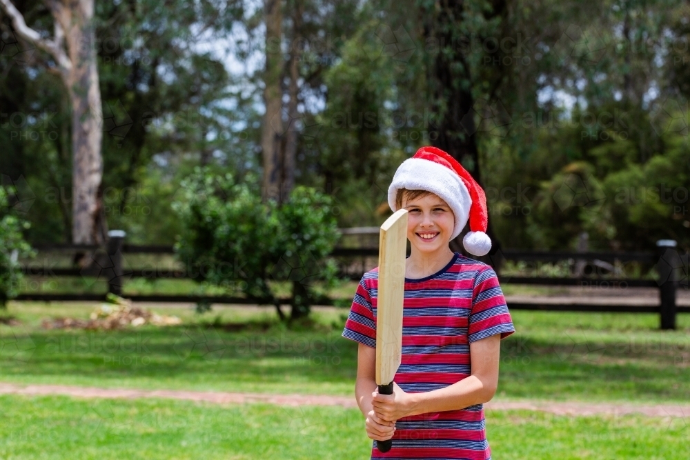 Aussie boy ready to play backyard cricket in front yard at Christmastime wearing santa hat - Australian Stock Image