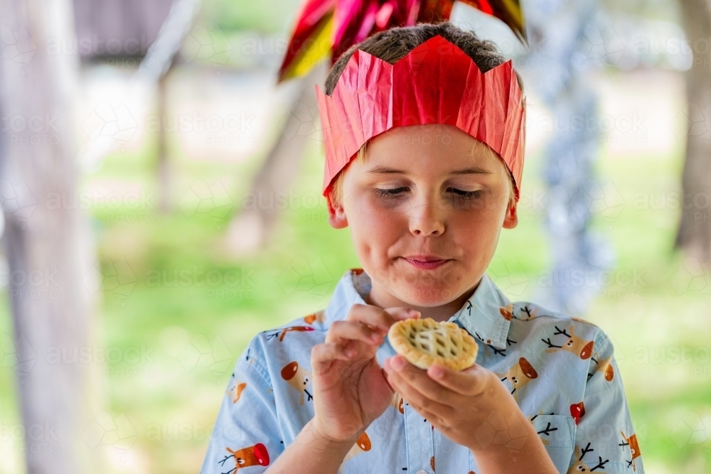 Aussie child staring hungrily at fruit mince pie in hands while wearing paper crown at Christmas - Australian Stock Image