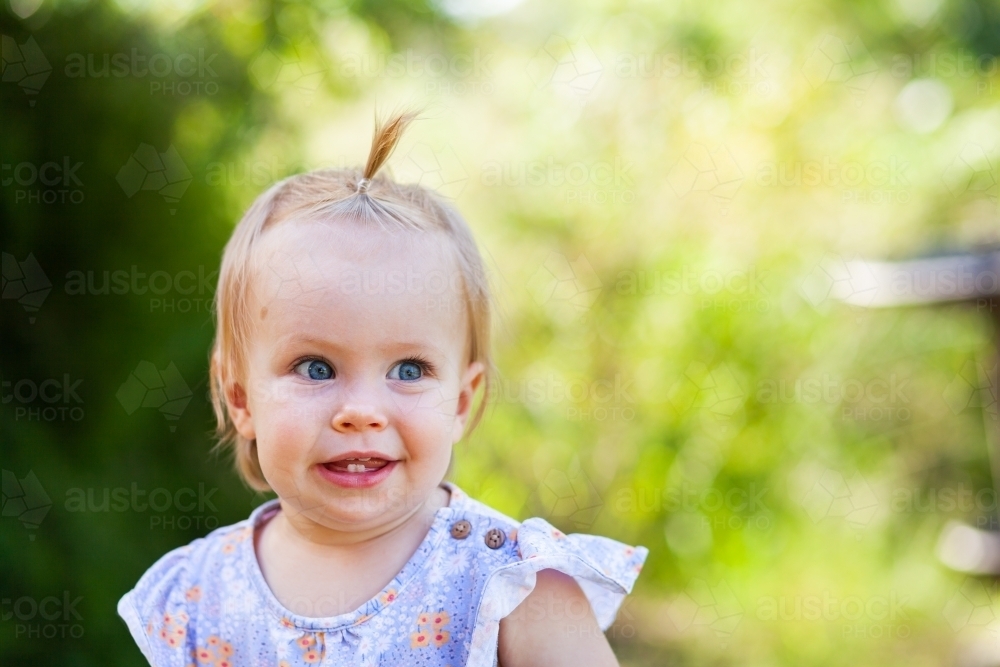 Baby girl against green backdrop outside with hair in topknot crest - Australian Stock Image