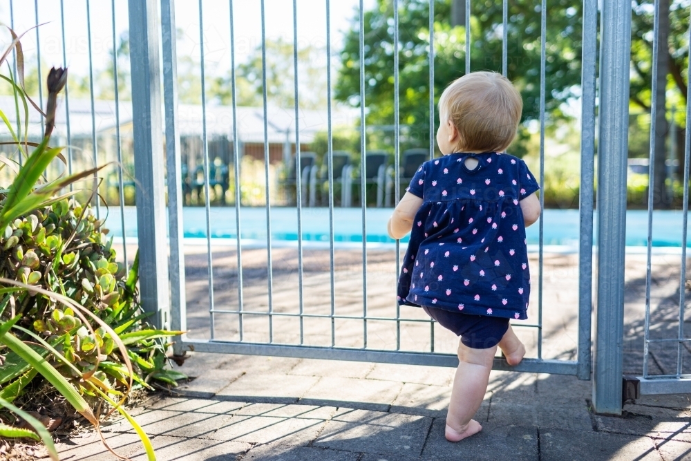 Baby girl at pool gate watching people splashing in the pool - Australian Stock Image