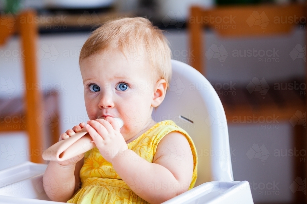 Baby girl in high chair in kitchen with milk ice block to keep cool in summer - Australian Stock Image