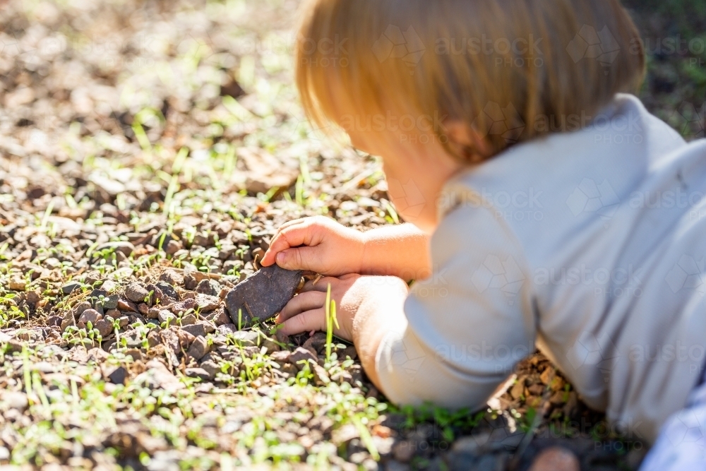 Baby girl lying on ground in gravel playing with rocks - Australian Stock Image