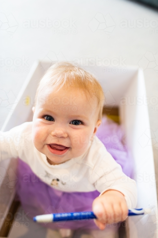 Baby girl sitting in box drawing with crayon textas at home - Australian Stock Image