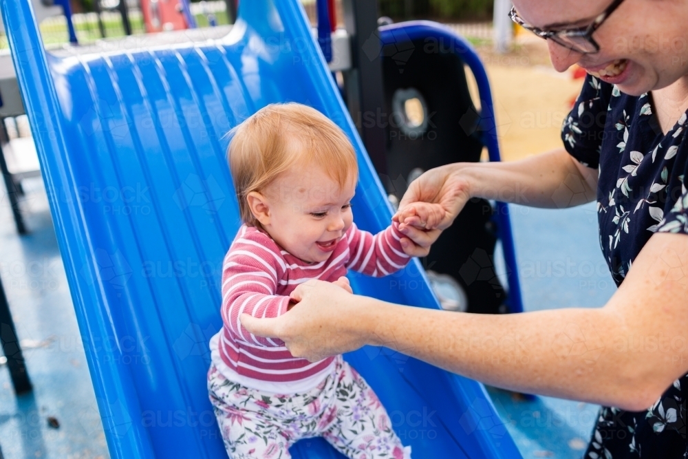 Baby holding mothers hands to slide down slippery dip at playground - Australian Stock Image