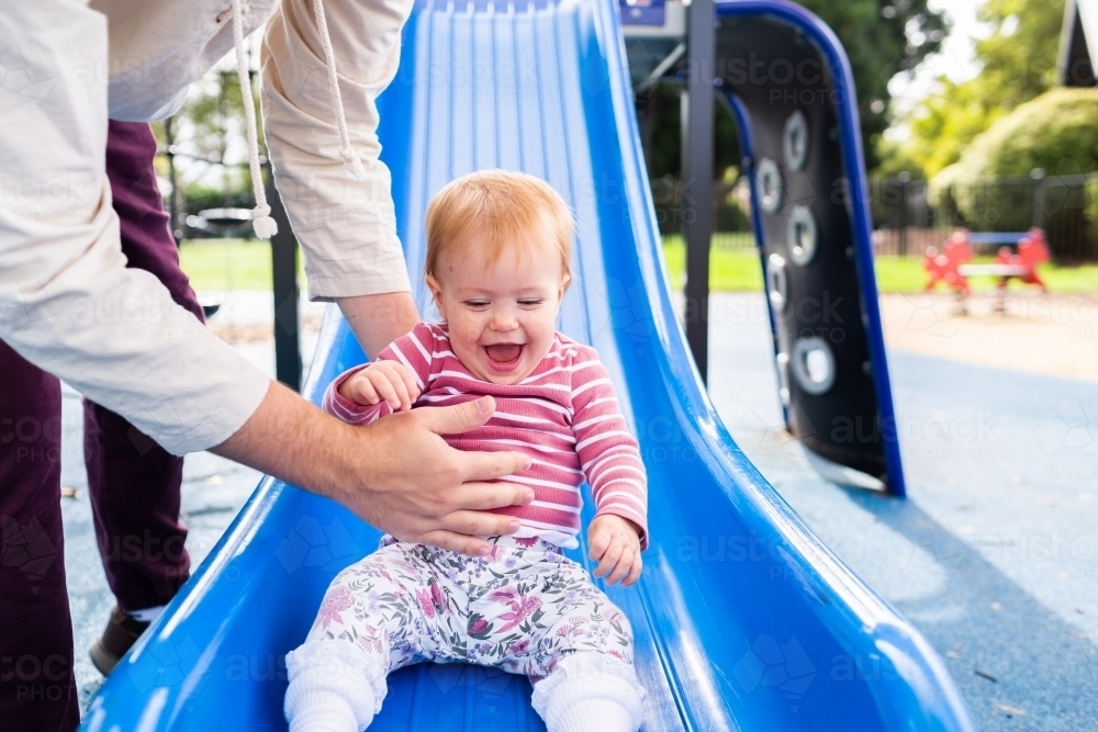 Baby laughing and being caught by father at playground slippery dip - Australian Stock Image