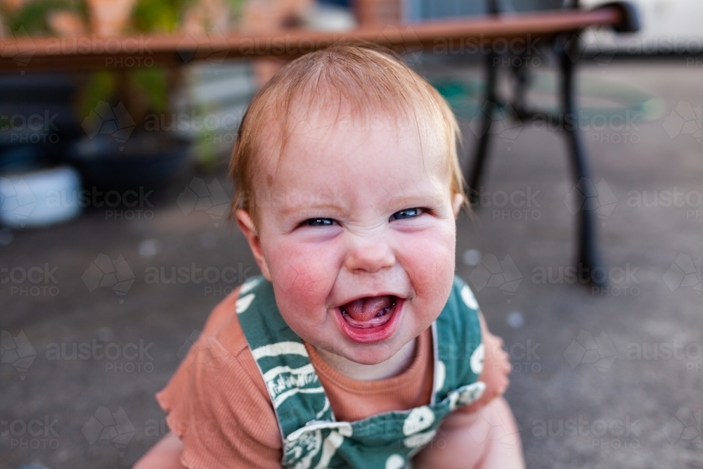 Baby laughing and pulling face outside playing in overalls - Australian Stock Image