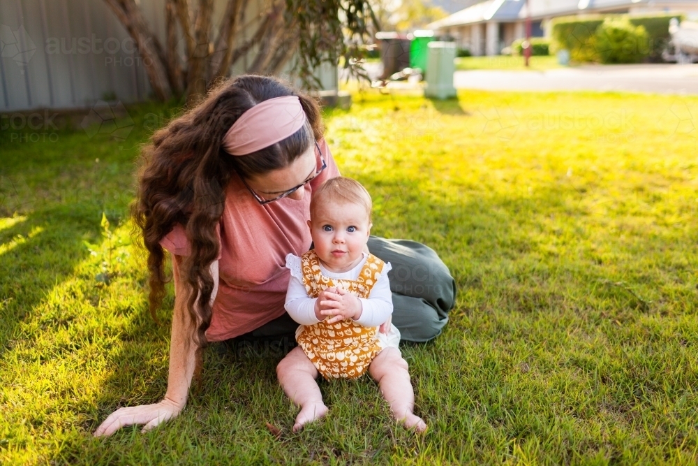 Baby looking at camera close to mum sitting on grass in front yard - Australian Stock Image