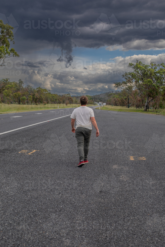 Back of a boy walking away along a road underneath a stormy sky - Australian Stock Image