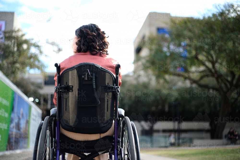 Back shot of a woman with disability sitting in wheelchair outside with trees around her - Australian Stock Image