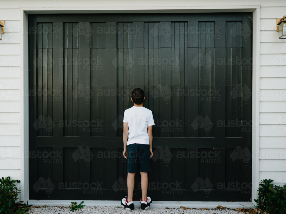 Back view of a young boy standing in front of a dark garage door. - Australian Stock Image