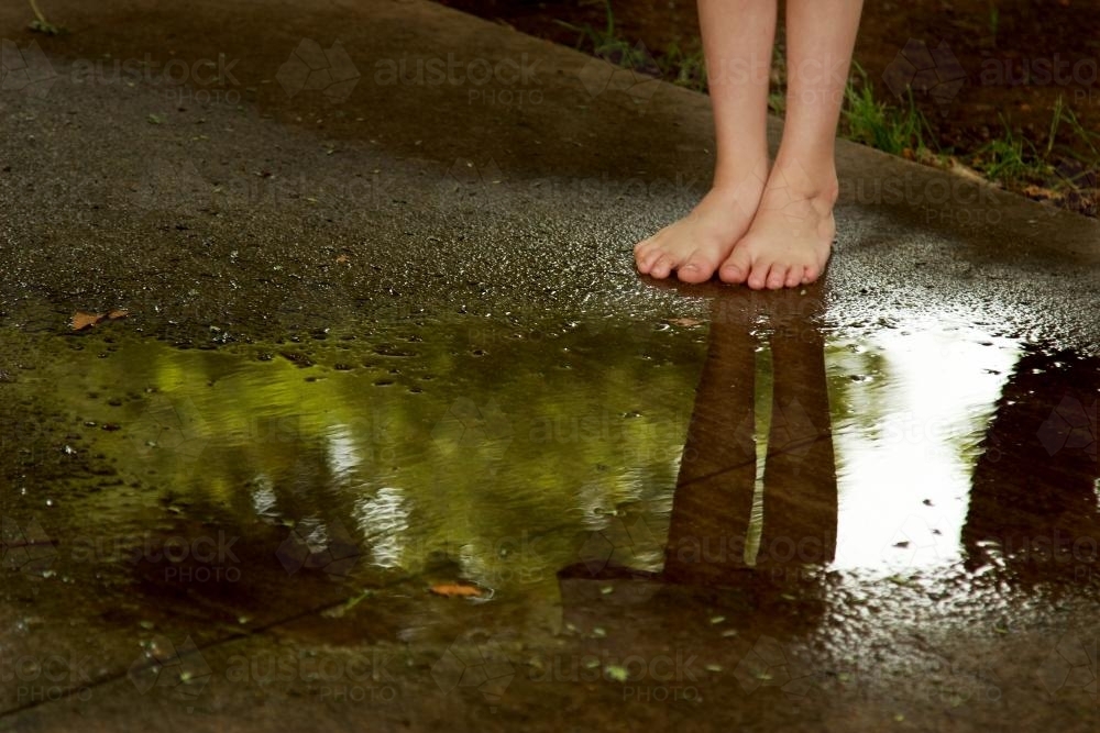 Bare feet of a young child reflected in a puddle on the footpath - Australian Stock Image