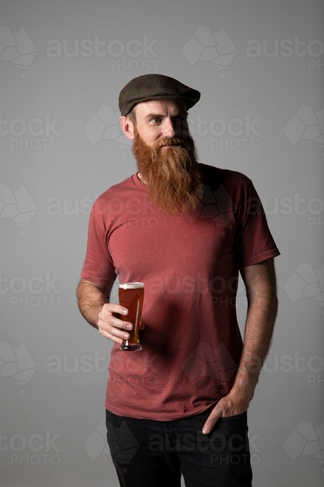 Bearded man in tweed flat cap standing holding a beer. - Australian Stock Image