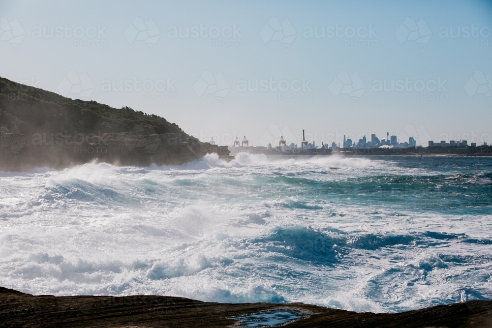 Big ocean swells along coast with cityscape in background - Australian Stock Image