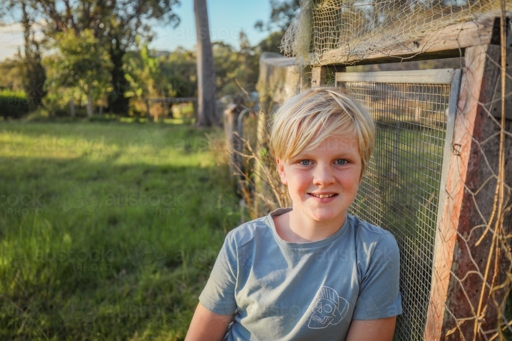 Blonde 10 year old boy relaxed and smiling on farm leaning against chicken coop - Australian Stock Image