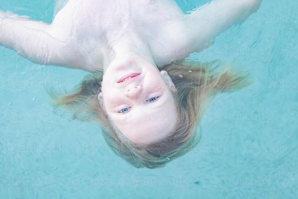 Blonde boy with long hair floating in pool with bright blue eyes - Australian Stock Image