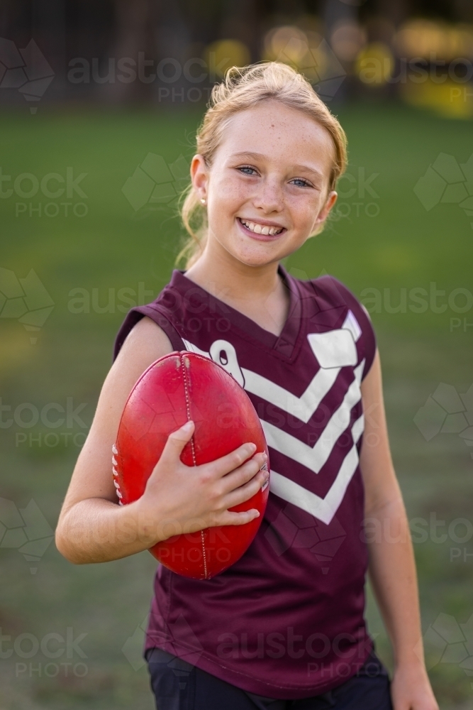 blonde girl holding red leather football and wearing football uniform - Australian Stock Image