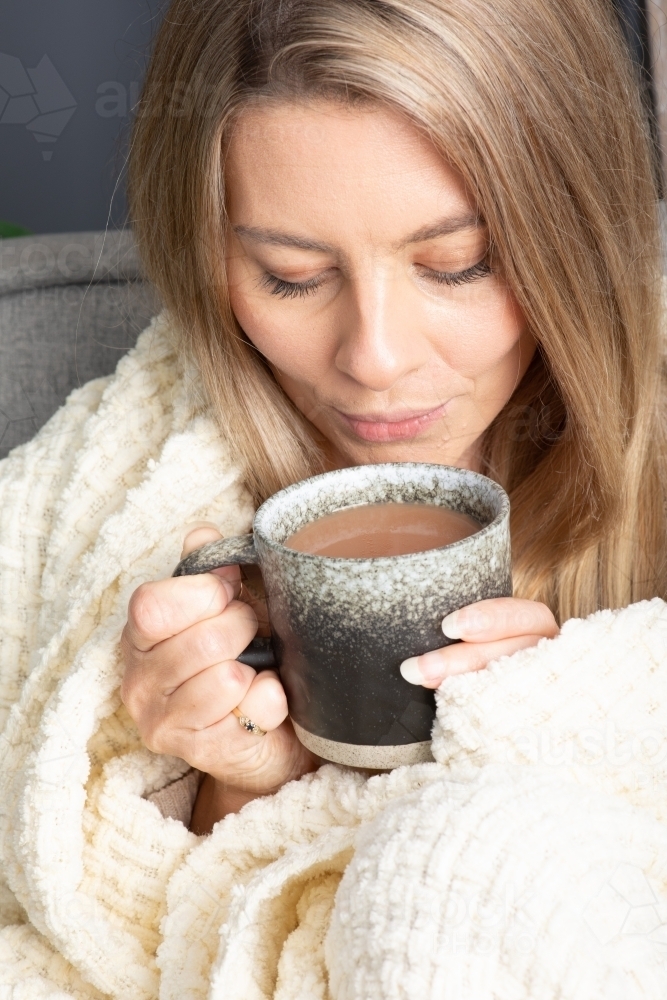 Blonde young lady wrapped in cream blanket hugging a mug - Australian Stock Image