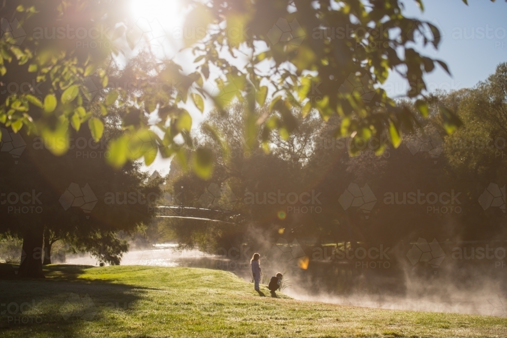 Boy and girl rugged up on cold morning fog day watching ducks in river - Australian Stock Image