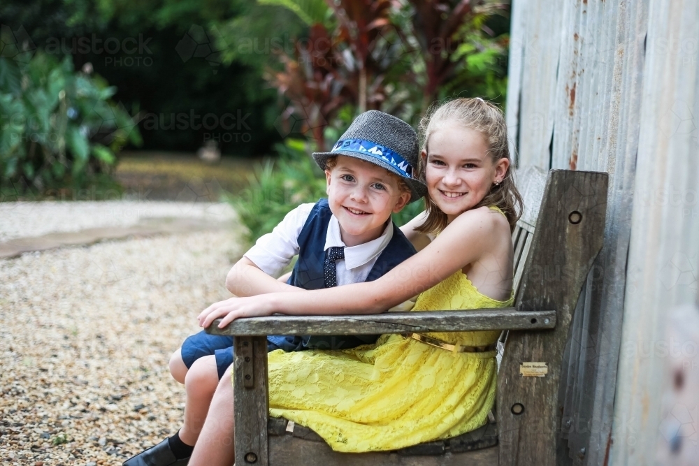 Boy and girl sitting on wooden bench seat smiling - Australian Stock Image