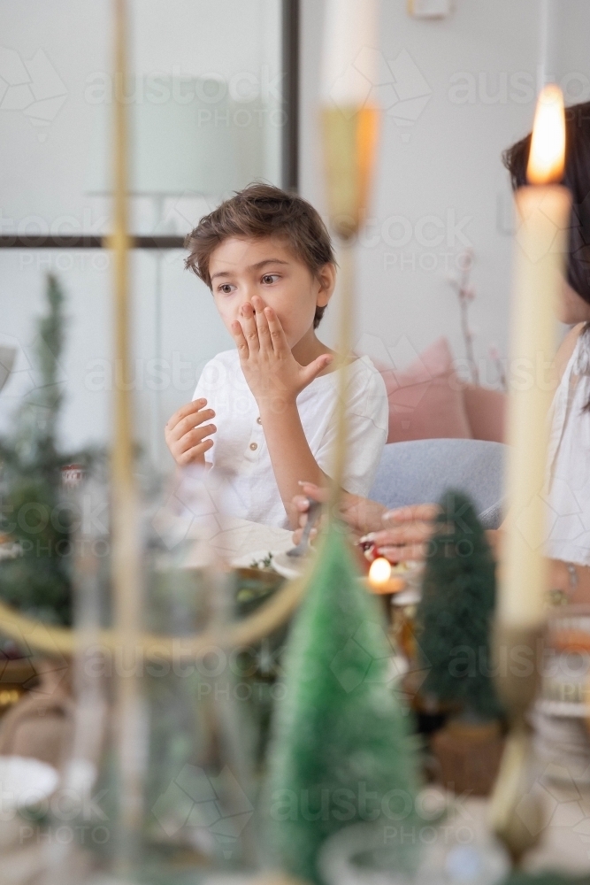 Boy covering his mouth while eating with festive decorated table in foreground - Australian Stock Image