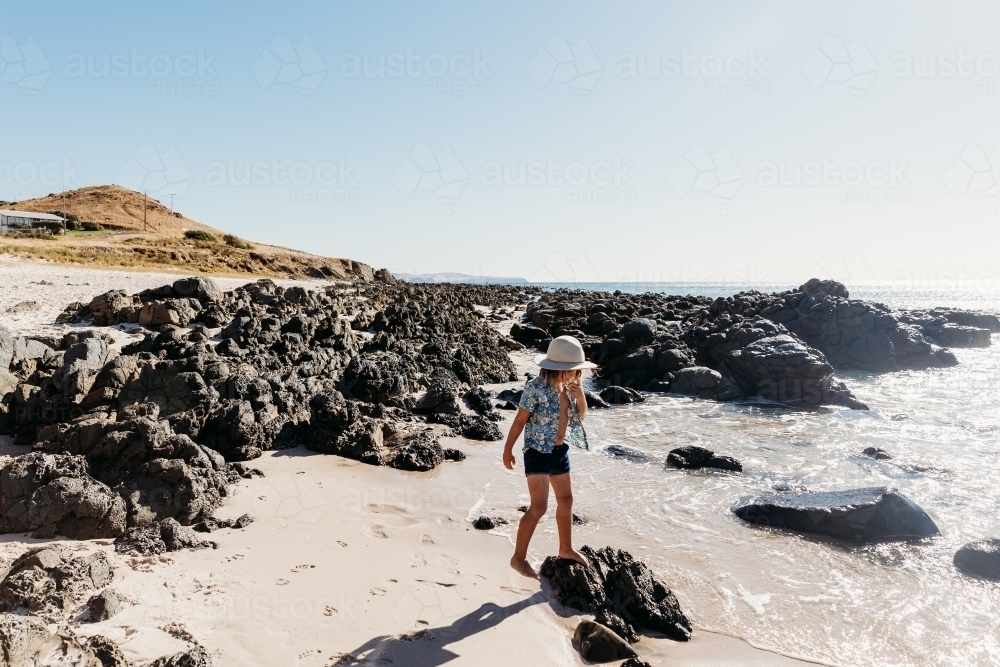 Boy in hat walking along beach with rocks and rolling hills behind him - Australian Stock Image