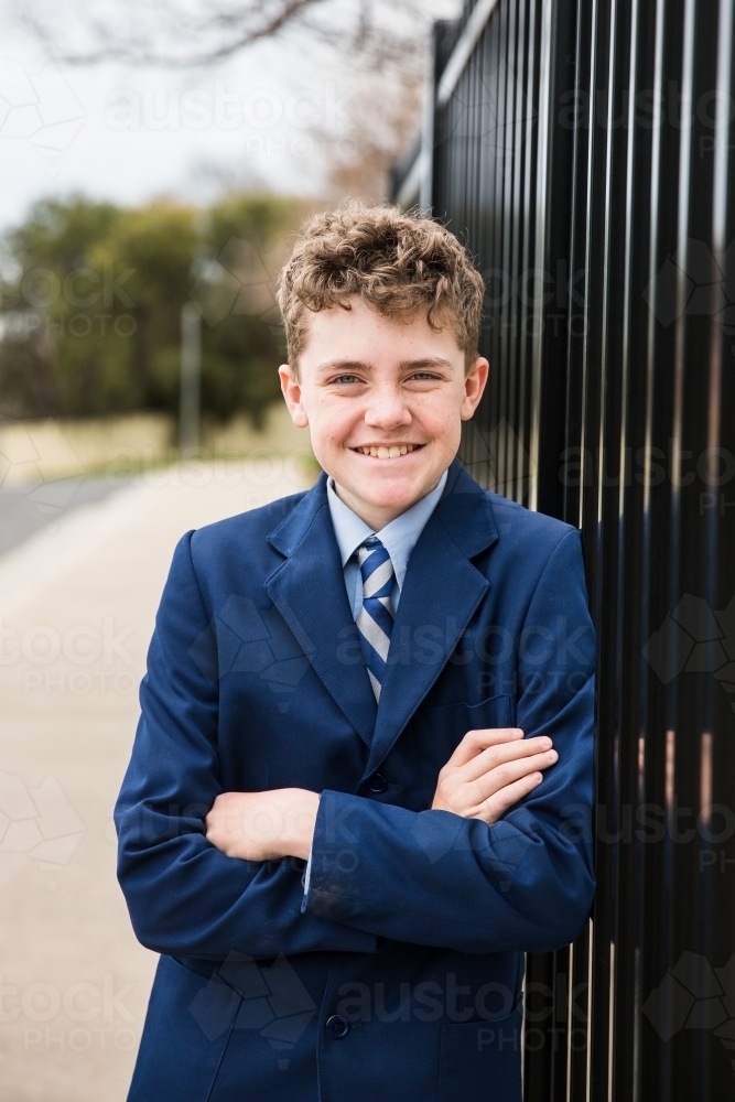 Boy in school uniform smiling with arms crossed leaning against fence at front of school - Australian Stock Image