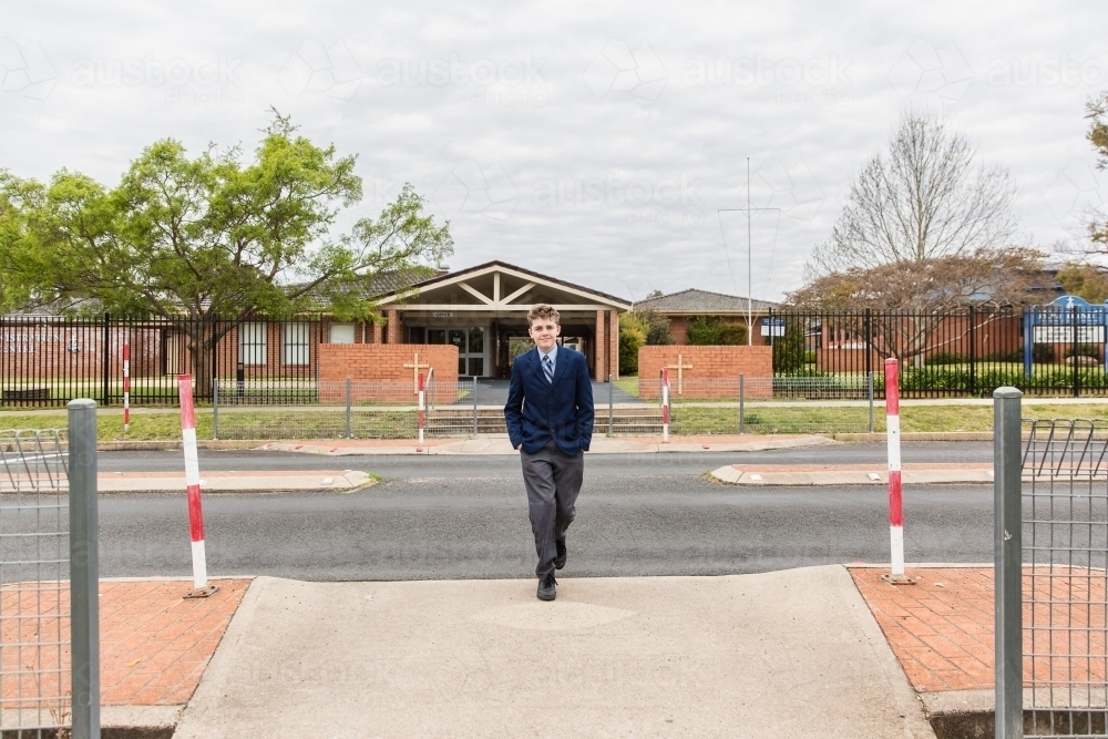 Boy in uniform crossing road in front of school with hands in pockets - Australian Stock Image