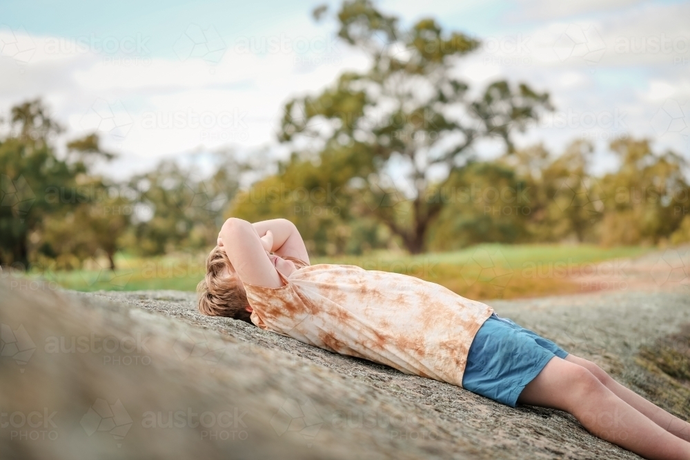 Boy lying outdoors on rock covering face from sun - Australian Stock Image