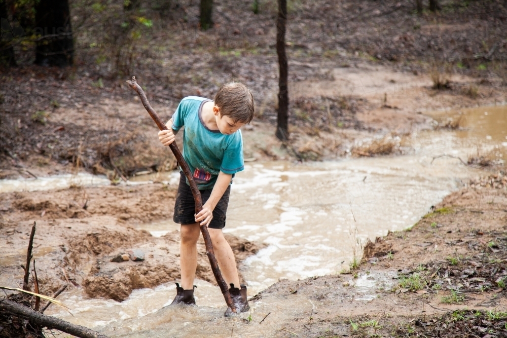 Boy playing in muddy creek with brown flowing water - Australian Stock Image