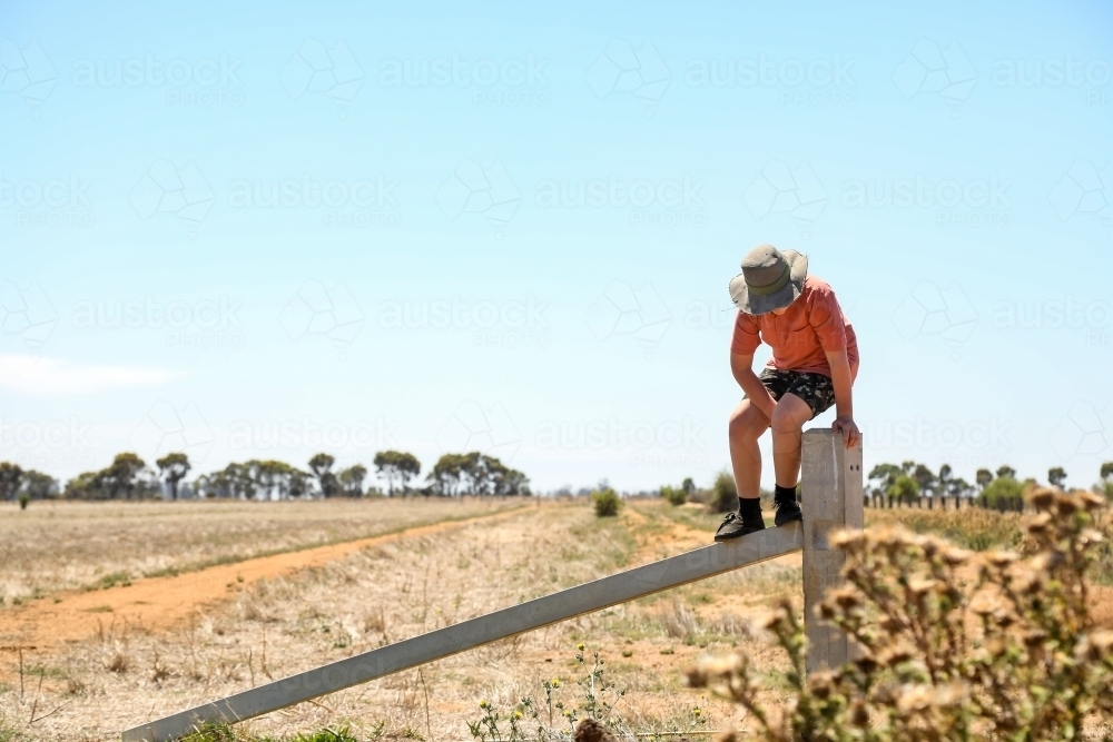 Boy sitting on farm fence in dry summer conditions - Australian Stock Image