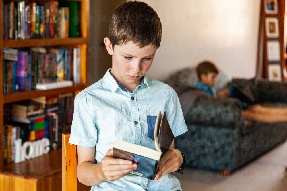 Boy sitting on kitchen stool reading a book - Australian Stock Image