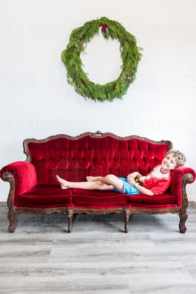 Boy sitting on red lounge smiling at Christmas with wreath - Australian Stock Image