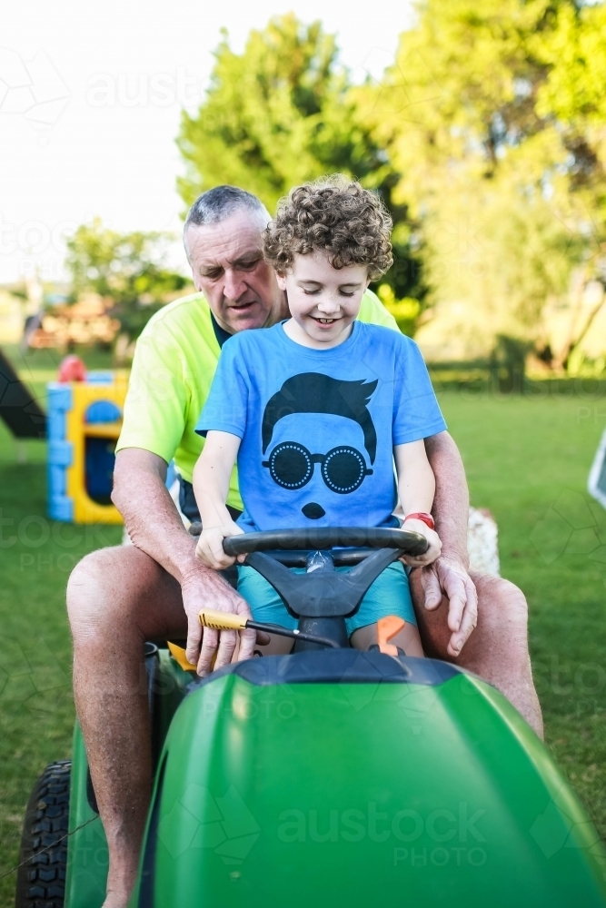 Boy sitting on ride on lawn mower with grandfather - Australian Stock Image