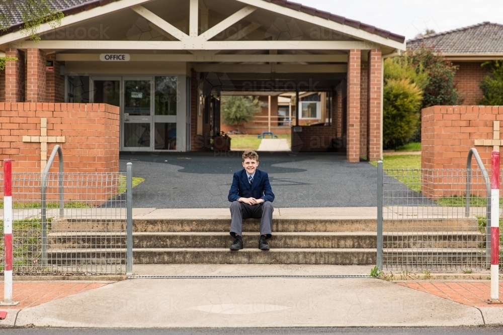 Boy sitting on steps at front of private catholic school smiling in uniform - Australian Stock Image