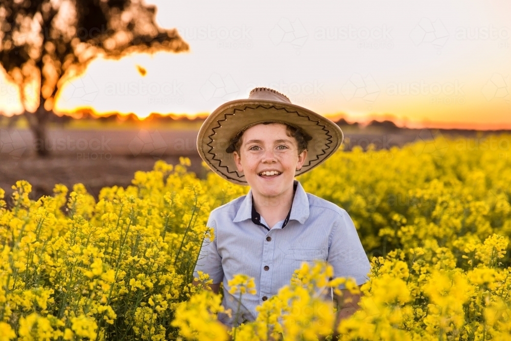 Boy smiling wearing hat in canola paddock at sunset on farm - Australian Stock Image