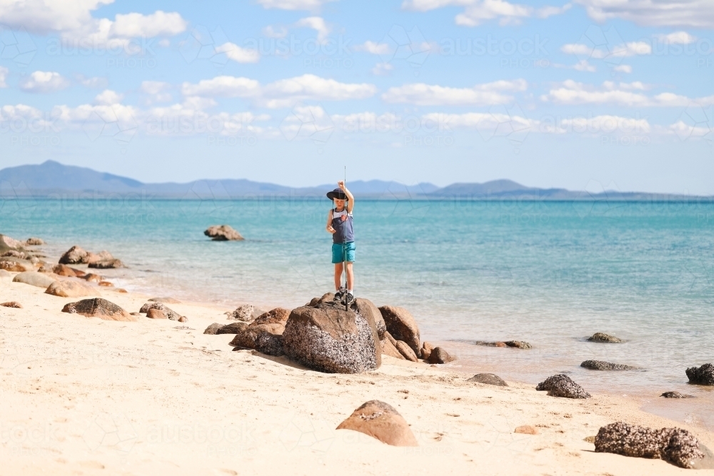 Boy standing on rocks with fishing rod on tropical beach with islands in the distance - Australian Stock Image