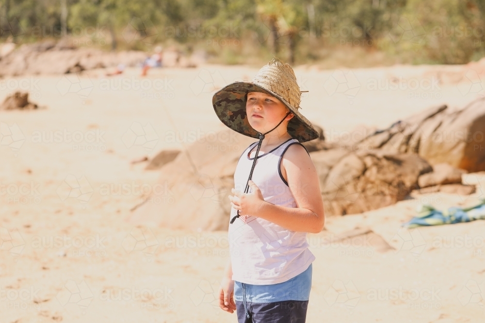 Boy standing on the beach wearing large straw hat at Cape Gloucester in the Whitsundays - Australian Stock Image