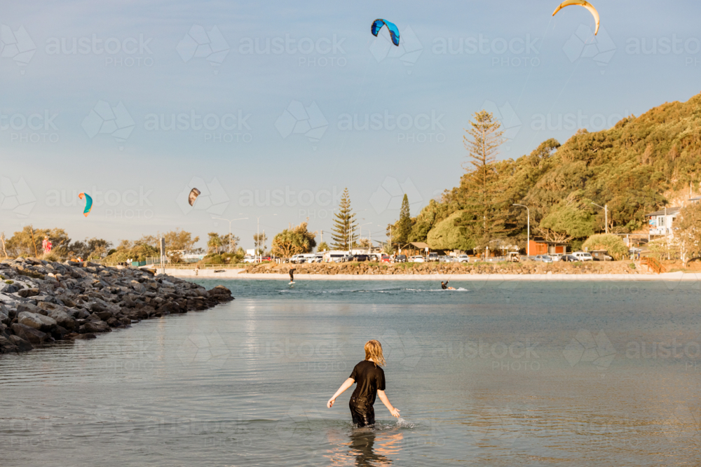 Boy wading in shallow water of Currumbin Creek watching kite surfers in the distance - Australian Stock Image