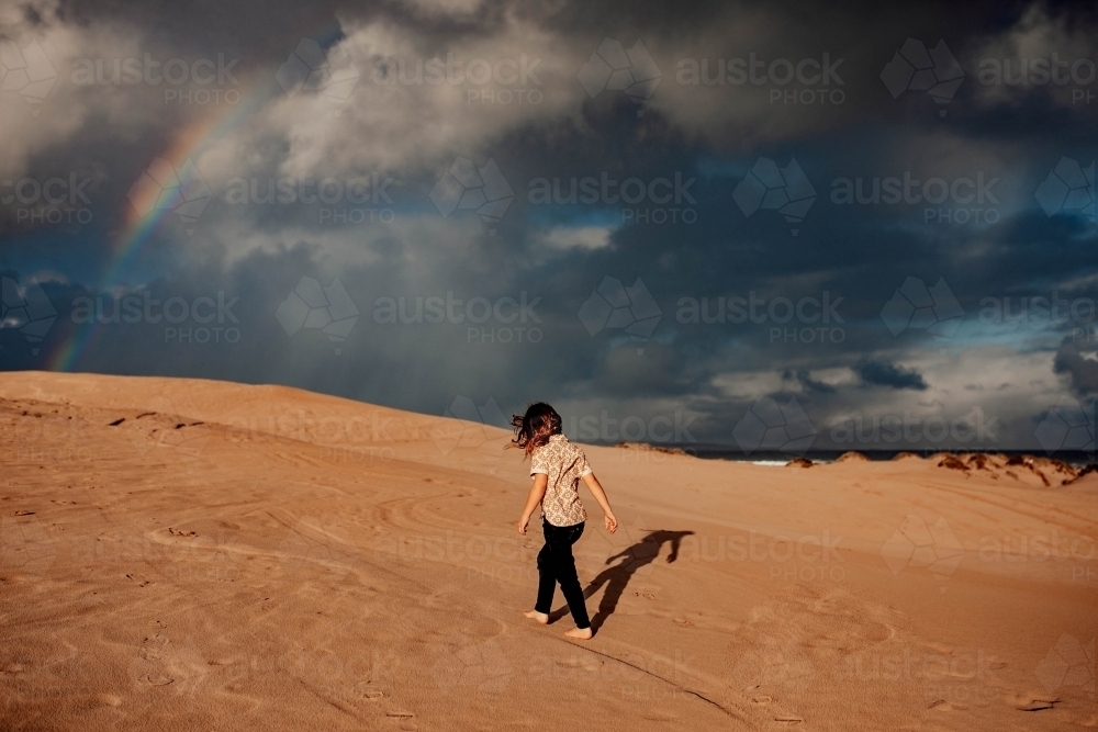 Boy walking on sand dune with rainbow in sky - Australian Stock Image