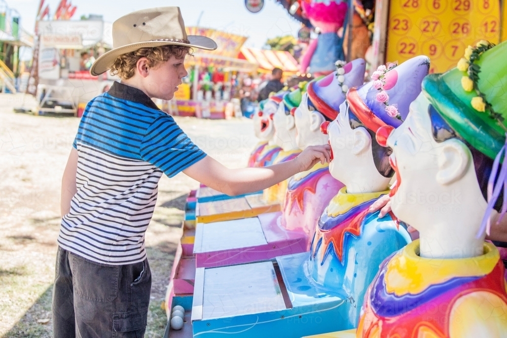 Boy wearing akubra hat placing ball in mouth of clowns sideshow alley at local show - Australian Stock Image