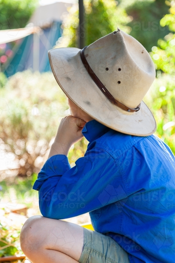 Boy wearing akubra hat squatting outside facing away - Australian Stock Image