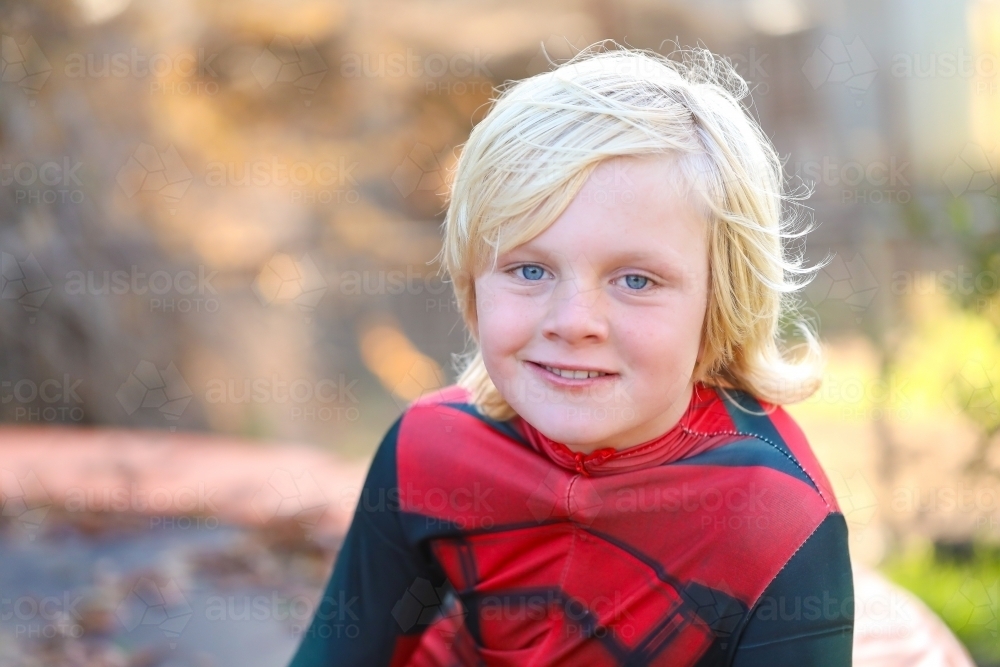 Boy wearing red superhero suit sitting on trampoline covered in autumn leaves - Australian Stock Image