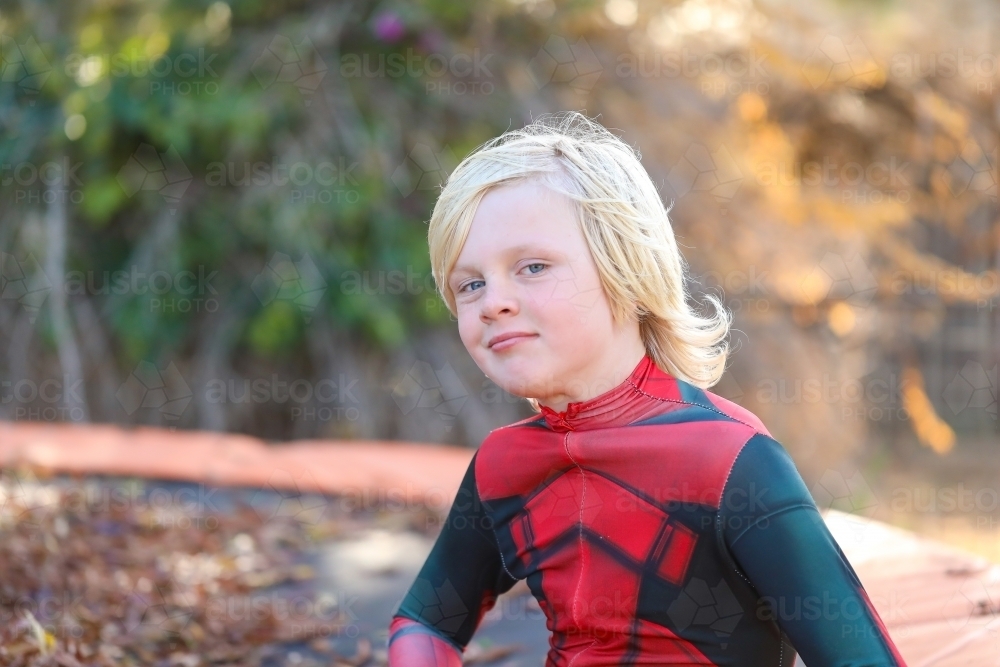 Boy wearing red superhero suit sitting on trampoline covered in autumn leaves - Australian Stock Image