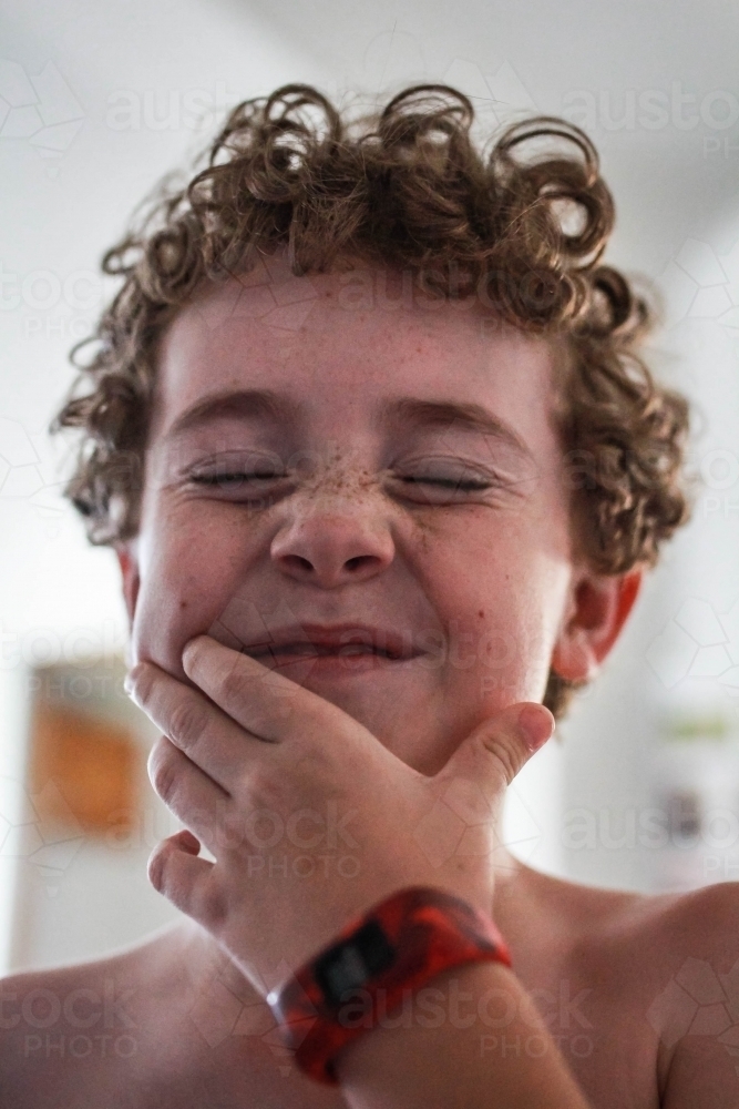 Boy with curly hair squinting eyes shut laughing with hand up to mouth - Australian Stock Image