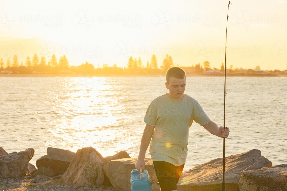 Boy with fishing equipment on the Stockton breakwall in golden afternoon light - Australian Stock Image